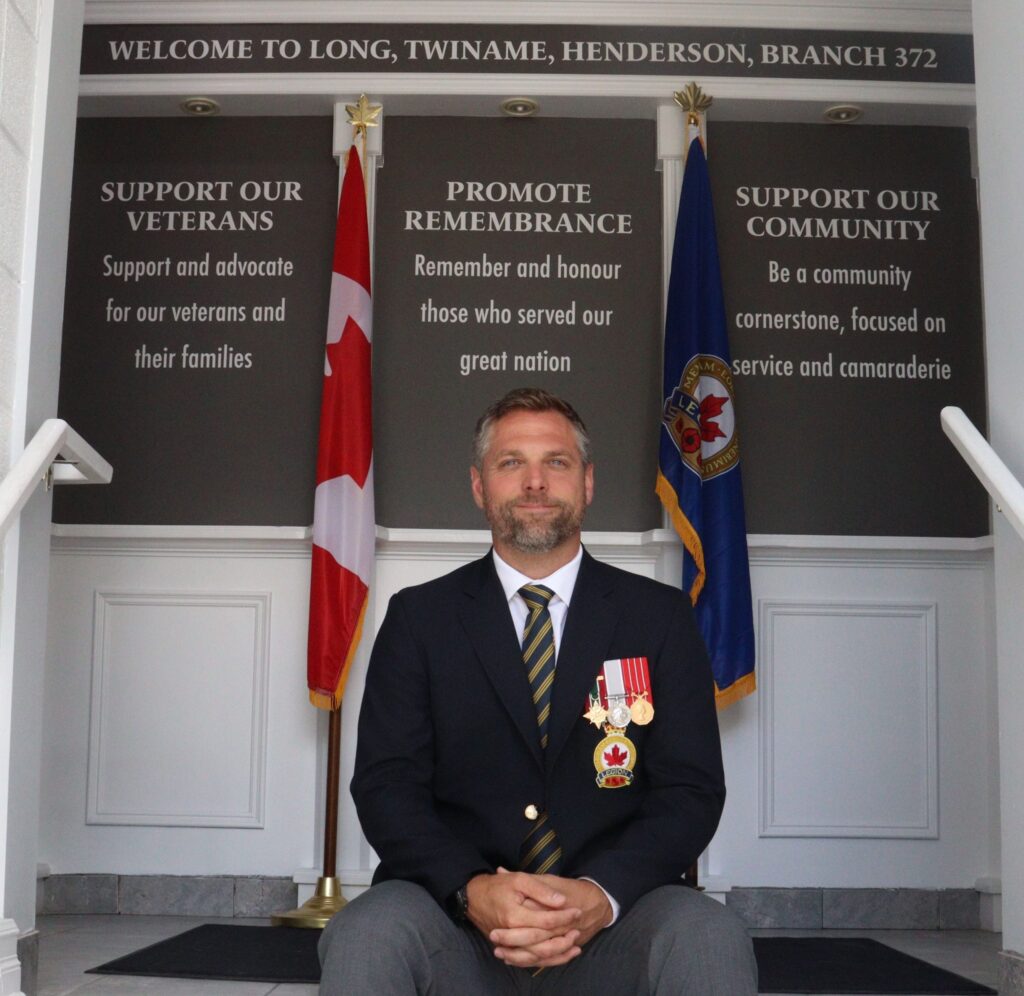 Paul Brown, president of the Russell Legion, sits beneath the organization’s three core values: supporting veterans, promoting remembrance, and serving the community, which are displayed at the entrance of the newly reopened branch.